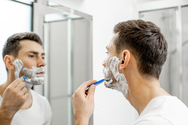 HANDSOME MAN IN FRON OF VANITY MIRROR WITH RAZOR IN HAND AND SHAVING CREAM ON FACE, SHAVING