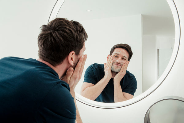 Man in dark t-shirt smiling while looking in vanity mirror and touching face